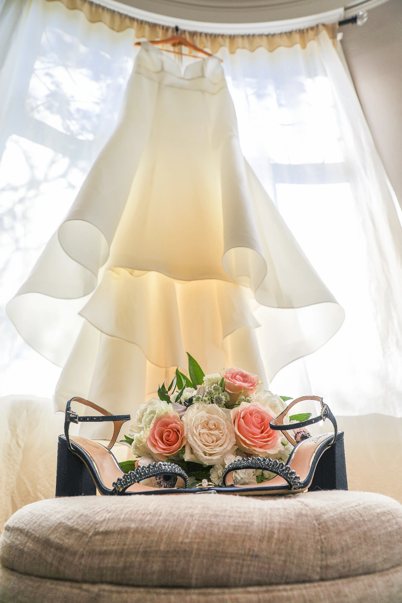 Bridal gown hanging in a sunlit window above a bouquet of blush roses and navy bridal heels